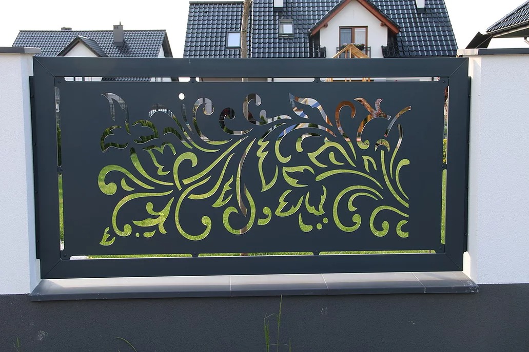 Nowoczesna, metalowa brama z dekoracyjnym, ażurowym wzorem roślinnym na tle zieleni ogrodu i fragmentu domu. Brama w kolorze grafitowym, osadzona na białych słupkach.