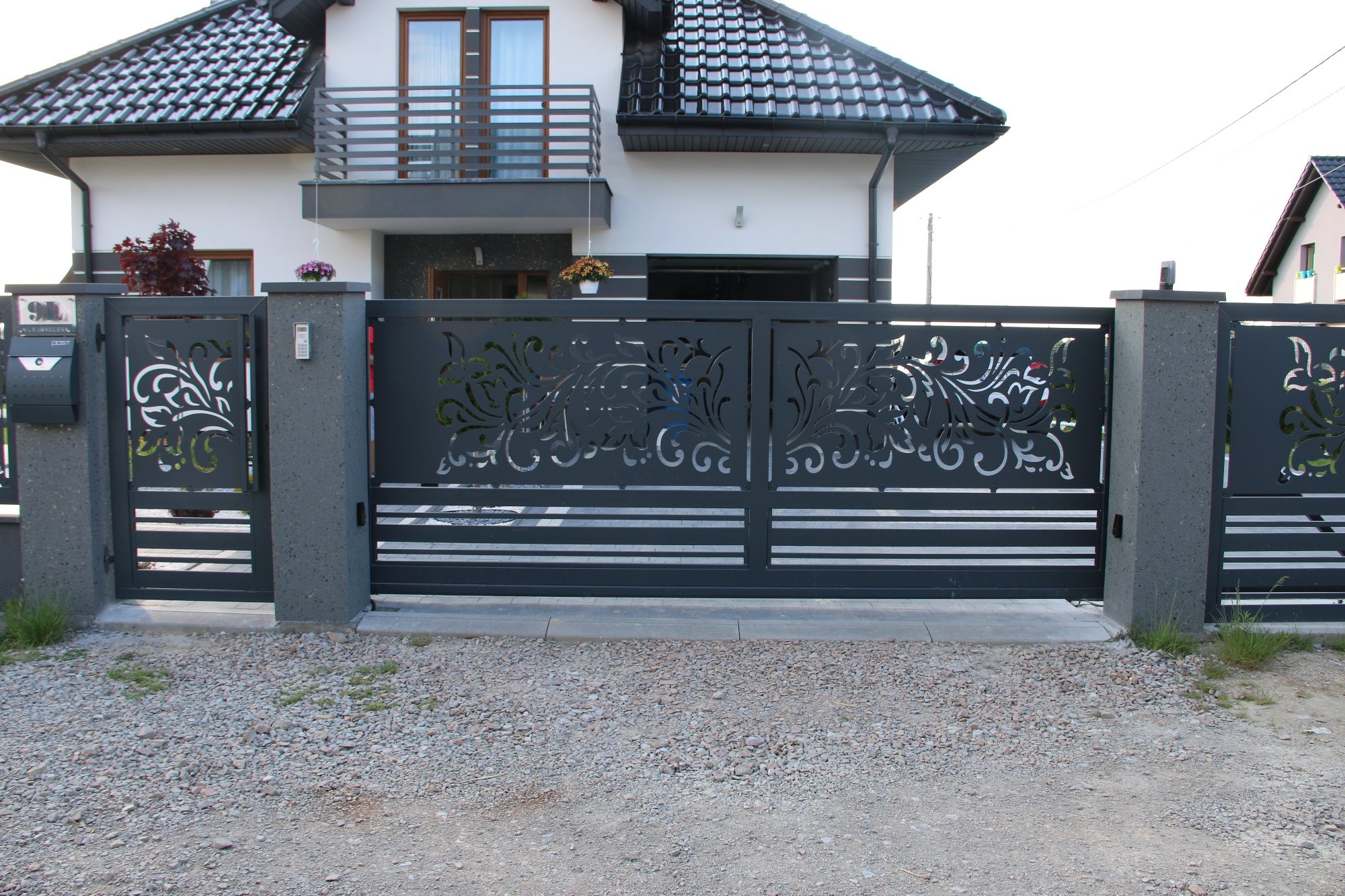 Nowoczesna, przesuwna brama wjazdowa z dekoracyjnym, ażurowym wzorem roślinnym, zamontowana przed domem z balkonem i szarą elewacją.
