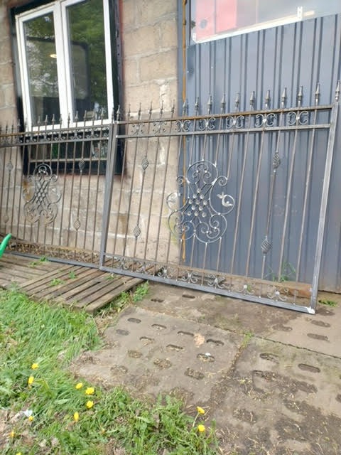 Dwa segmenty metalowego ogrodzenia z dekoracyjnymi elementami, oparte o ścianę i ziemię. Widoczne ślady spawania i surowy metal. Częściowo widoczny budynek w tle.