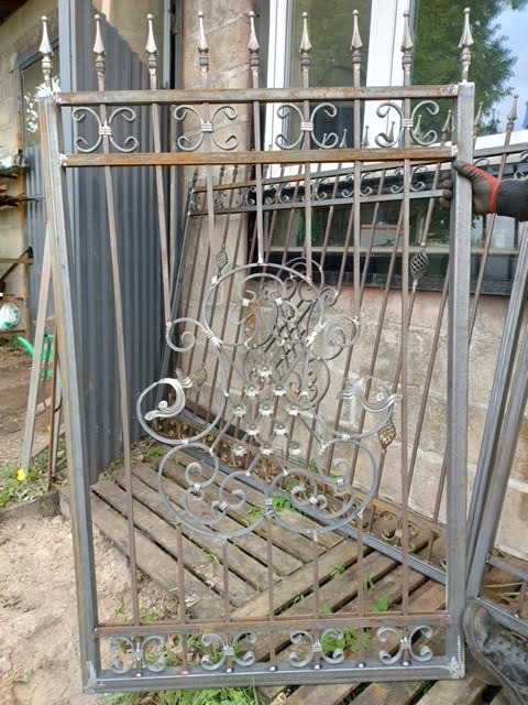 Metalowa brama z dekoracyjnymi elementami, częściowo oparta o paletę. Widoczna ręka w rękawicy trzymająca konstrukcję. Tło stanowią budynki i zieleń.