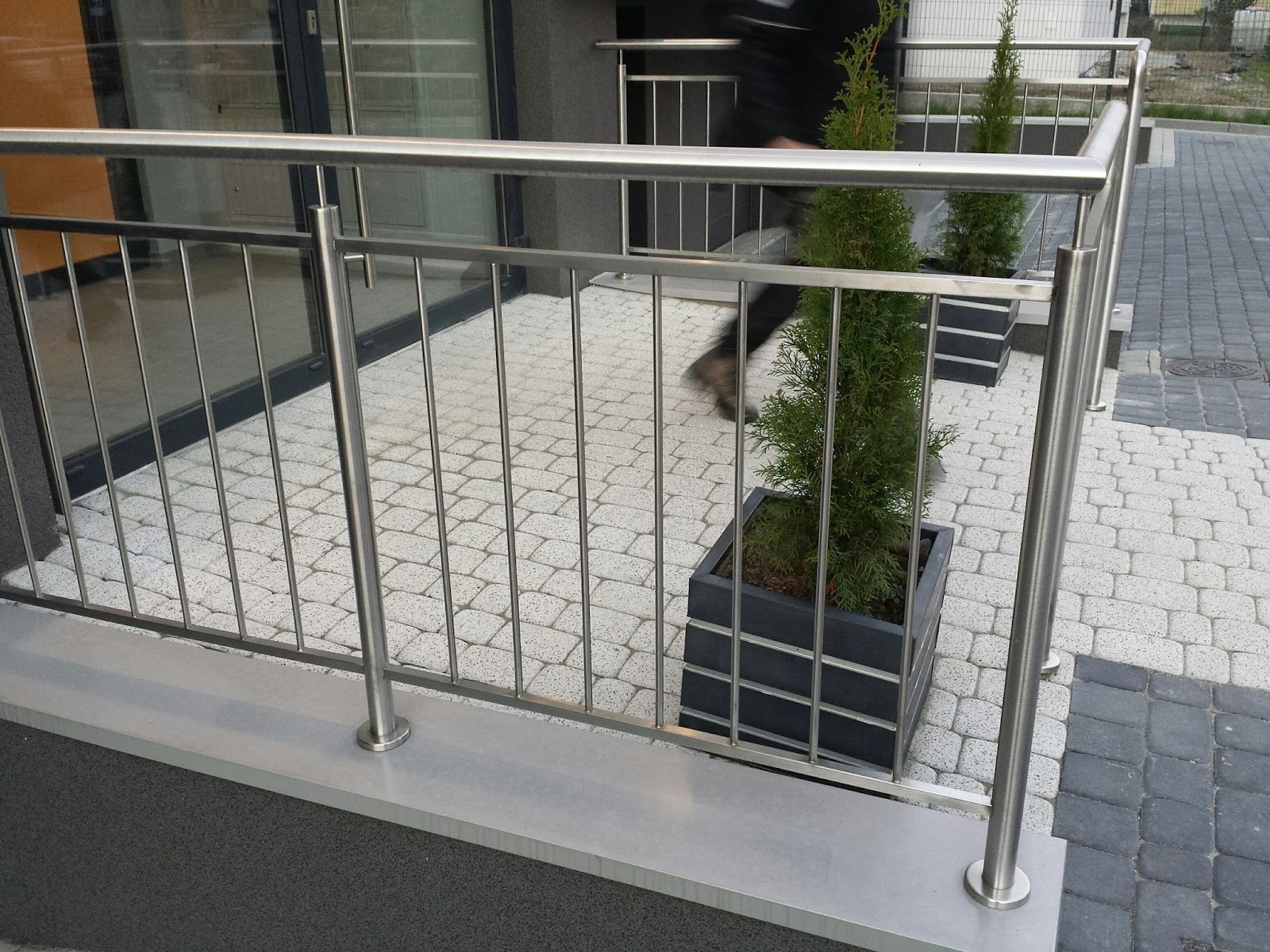 Nowoczesna balustrada ze stali nierdzewnej, z subtelnymi roślinami w donicach, na tle kostki brukowej. Widoczny fragment osoby przechodzącej obok.