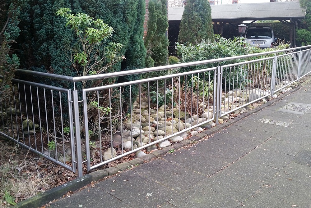 Nowoczesne ogrodzenie metalowe ze stali nierdzewnej, oddzielające posesję od chodnika. W tle roślinność i zadaszone miejsce parkingowe.