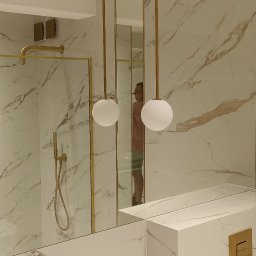 Expert Build - Nowoczesna łazienka z marmurowymi płytkami, złotymi akcentami i kulistymi lampami wiszącymi; widoczne kabina prysznicowa ze złotymi ramami i armaturą, umywalka z szafką oraz odbicie osoby w lustrze.
