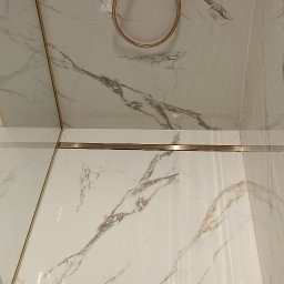 Expert Build - Białe płytki imitujące marmur z delikatnymi szarymi i brązowymi żyłkami, złota armatura prysznicowa i listwy wykończeniowe, widok z dołu.