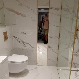 Expert Build - Nowoczesna łazienka z białymi płytkami imitującymi marmur, złotą armaturą i kabiną prysznicową typu walk-in, z odbiciem osoby w lustrze.