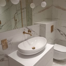 Expert Build - Nowoczesna łazienka z marmurowymi płytkami, owalną umywalką nablatową, złotą armaturą i lustrem z cienką złotą ramą, w której odbija się osoba robiąca zdjęcie.