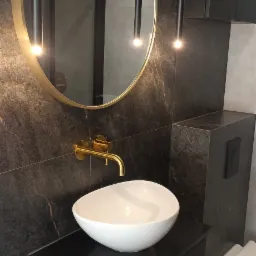 Nowoczesna łazienka z umywalką nablatową, złotą armaturą i okrągłym lustrem podświetlanym wiszącymi lampami, wykończona płytkami imitującymi ciemny marmur.