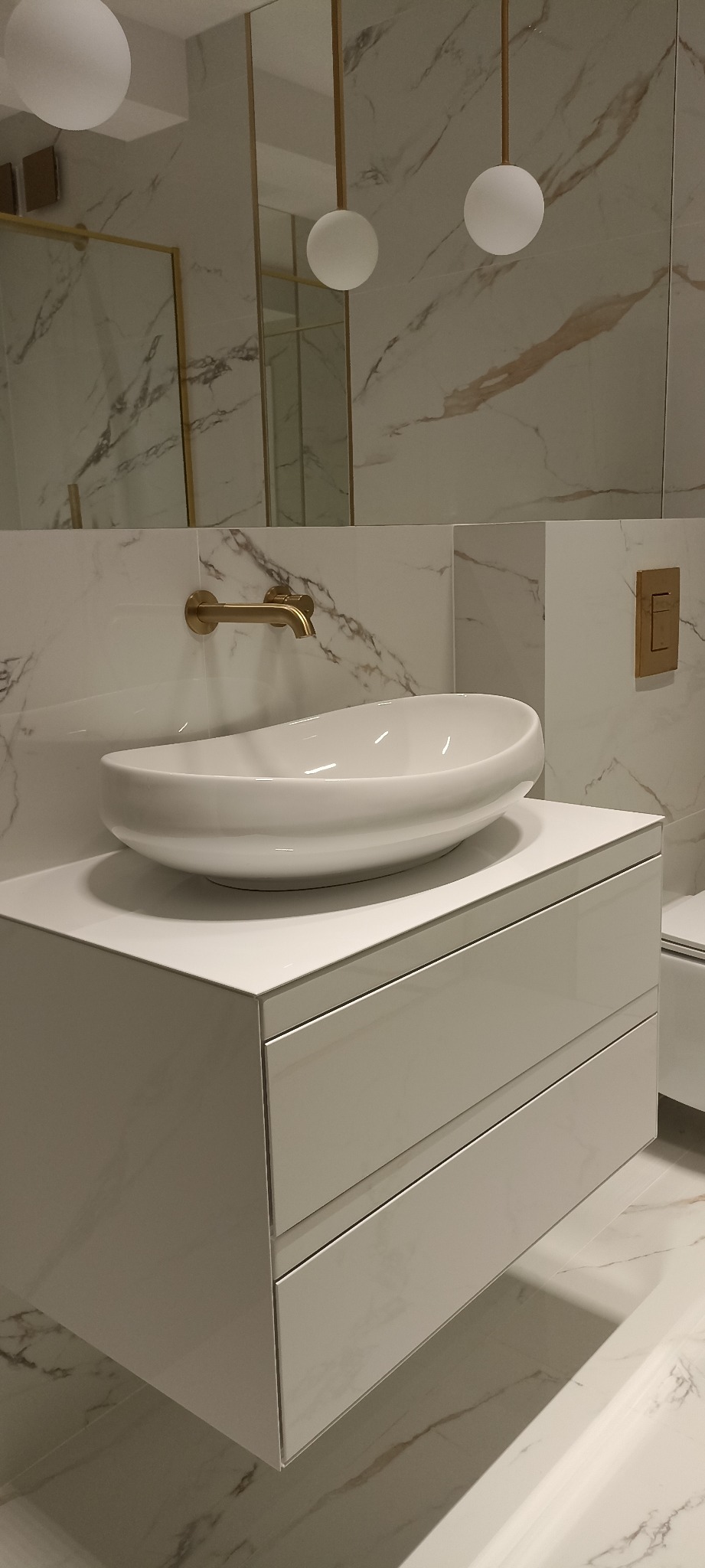 Nowoczesna łazienka z białą, owalną umywalką nablatową na szafce z dwiema szufladami, złota bateria ścienna, lustro w złotej ramie i marmurowe płytki na ścianach i podłodze, uzupełnione o kuliste...