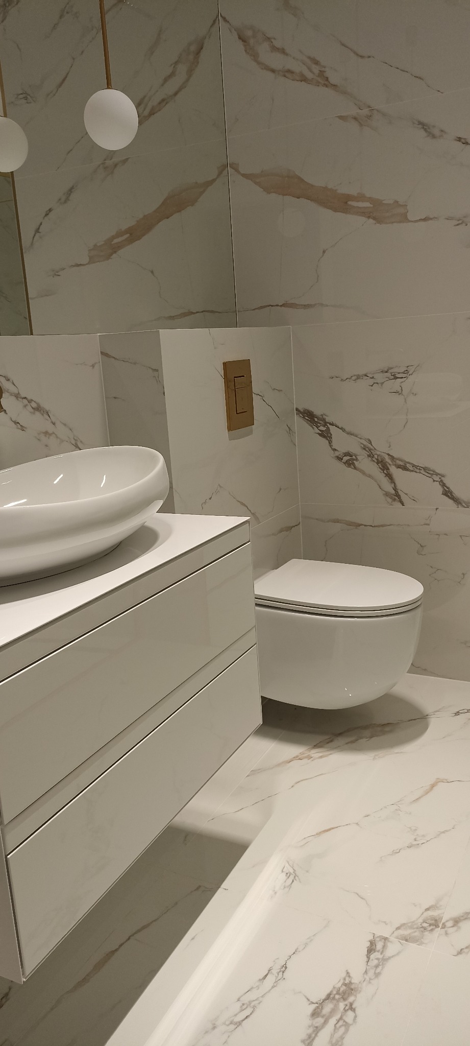 Nowoczesna łazienka z marmurowymi płytkami, wisząca toaleta, biała szafka pod umywalką z owalną, nablatową umywalką oraz złotym przyciskiem spłuczki.