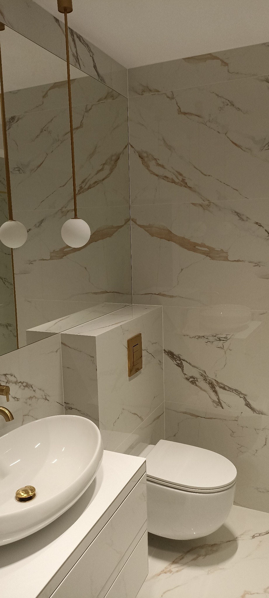 Nowoczesna łazienka z marmurowymi płytkami, wisząca toaleta, umywalka nablatowa z złotą armaturą i kuliste lampy wiszące.
