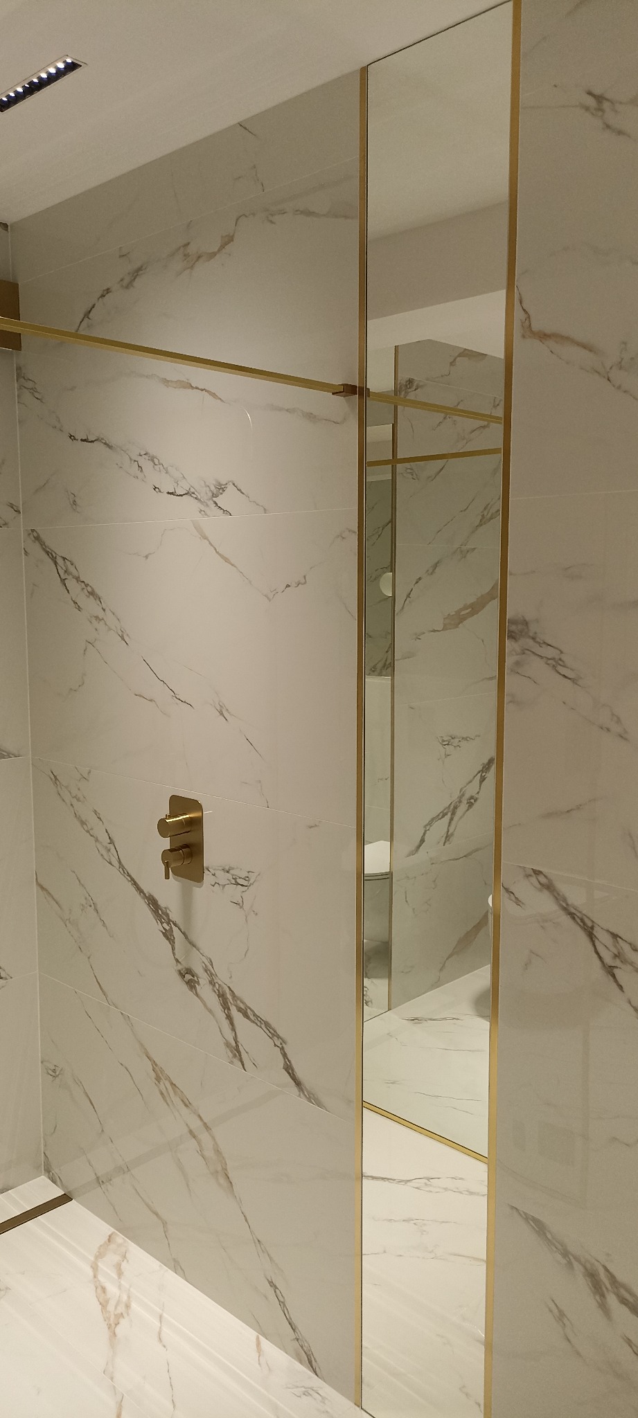 Nowoczesna łazienka z białymi płytkami imitującymi marmur, złotą armaturą i kabiną prysznicową ze złotymi profilami.