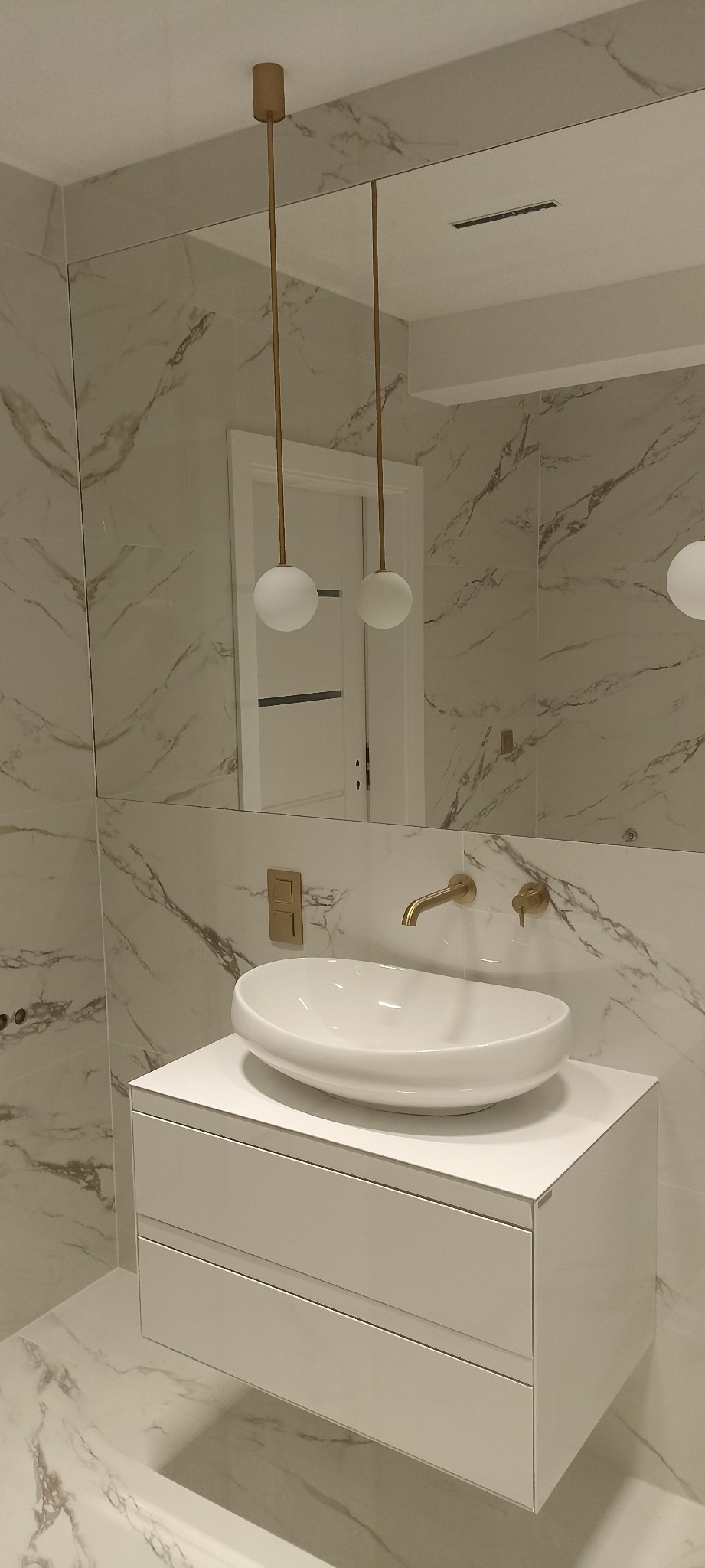 Nowoczesna łazienka z umywalką nablatową, złotą armaturą i marmurowymi płytkami, oświetlona wiszącymi lampami z kulistymi kloszami, odbicie białych drzwi w lustrze.