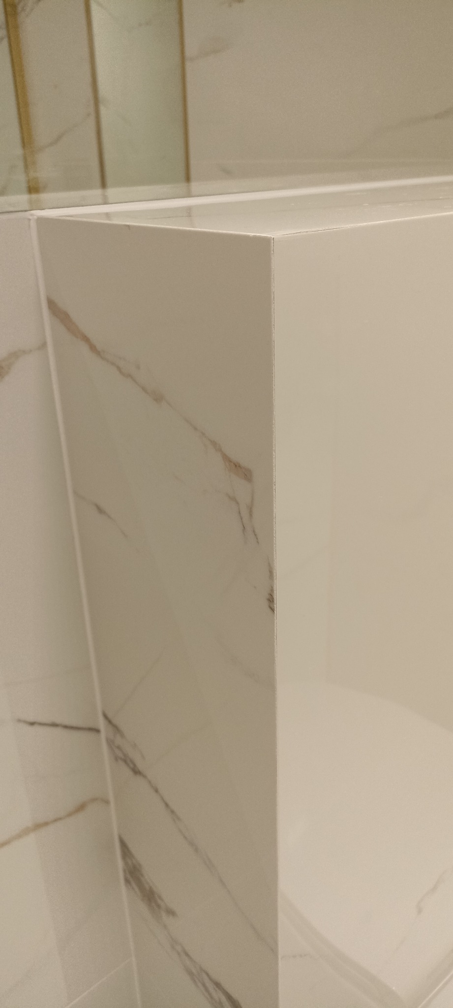 Fragment ściany wykończonej białymi płytkami imitującymi marmur, z widocznym narożnikiem i fragmentem gładkiej, białej powierzchni.