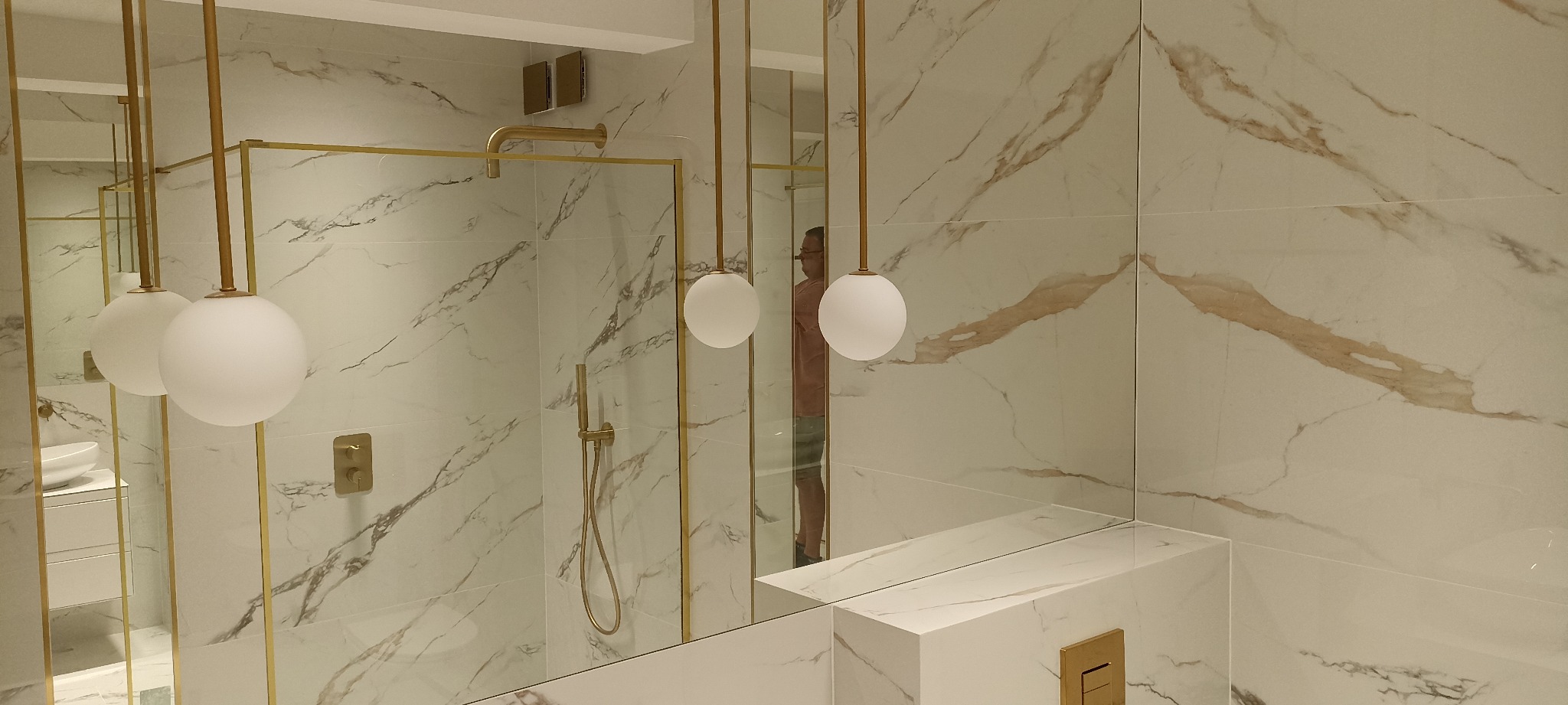 Nowoczesna łazienka z marmurowymi płytkami, złotymi akcentami i kulistymi lampami wiszącymi; widoczne kabina prysznicowa ze złotymi ramami i armaturą, umywalka z szafką oraz odbicie osoby w lustrze.