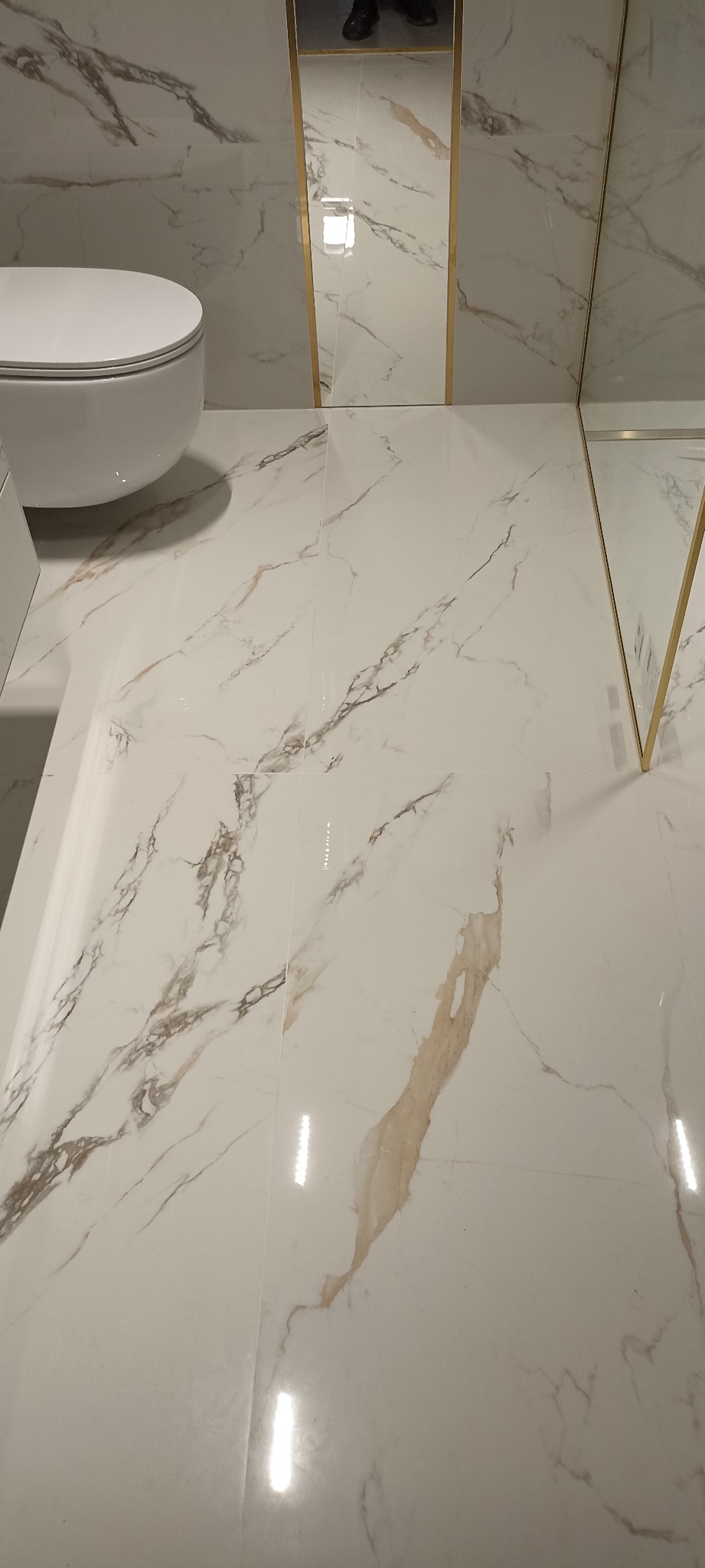 Białe płytki podłogowe i ścienne z marmurowym wzorem w łazience, z toaletą wiszącą i lustrem z cienką złotą ramą.