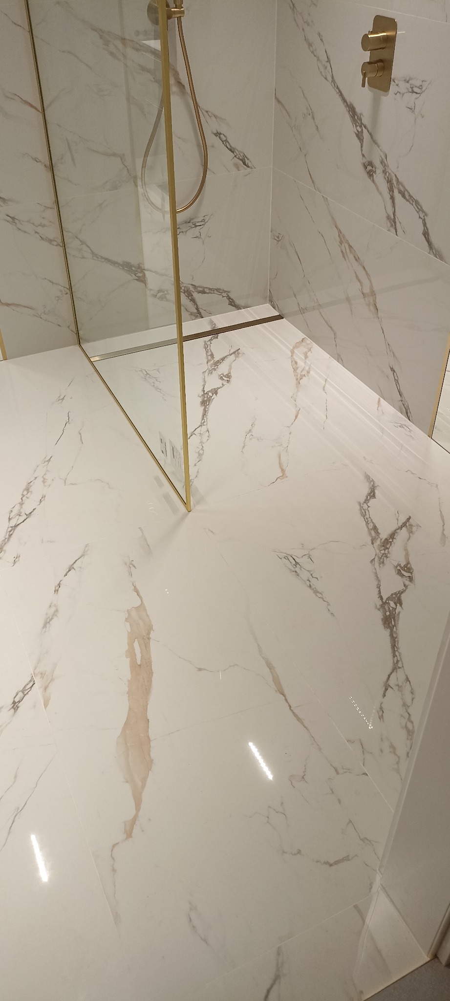 Nowoczesna kabina prysznicowa walk-in z białymi płytkami imitującymi marmur i złotymi detalami, takimi jak rama szyby i bateria.
