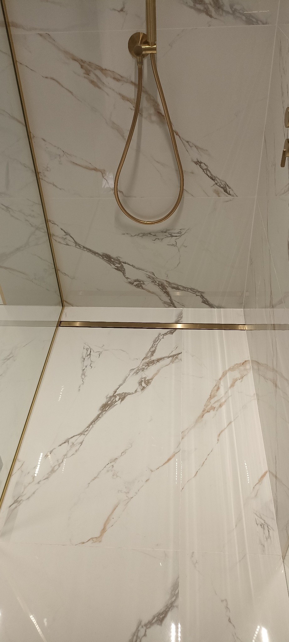 Białe płytki imitujące marmur z delikatnymi szarymi i brązowymi żyłkami, złota armatura prysznicowa i listwy wykończeniowe, widok z dołu.