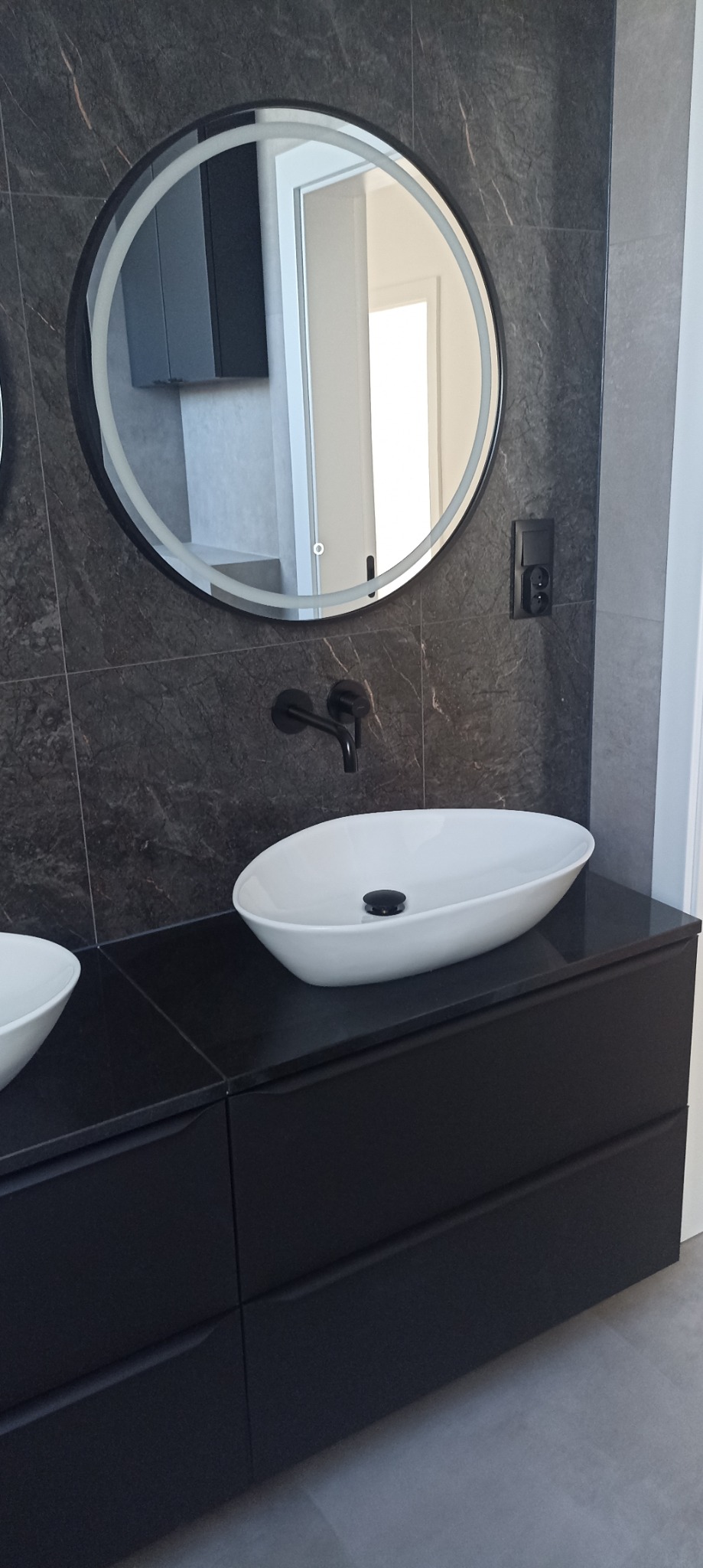 Nowoczesna łazienka z owalną białą umywalką nablatową, czarną armaturą i podświetlanym okrągłym lustrem na tle szarej ściany z płytek imitujących kamień.