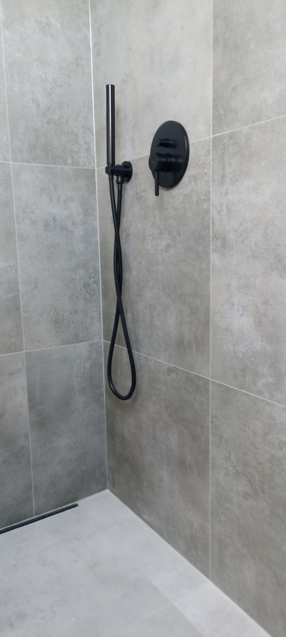 Szara łazienka z czarną armaturą: bateria natryskowa i deszczownica z wężem w nowoczesnym stylu, ułożone na płytkach imitujących beton.