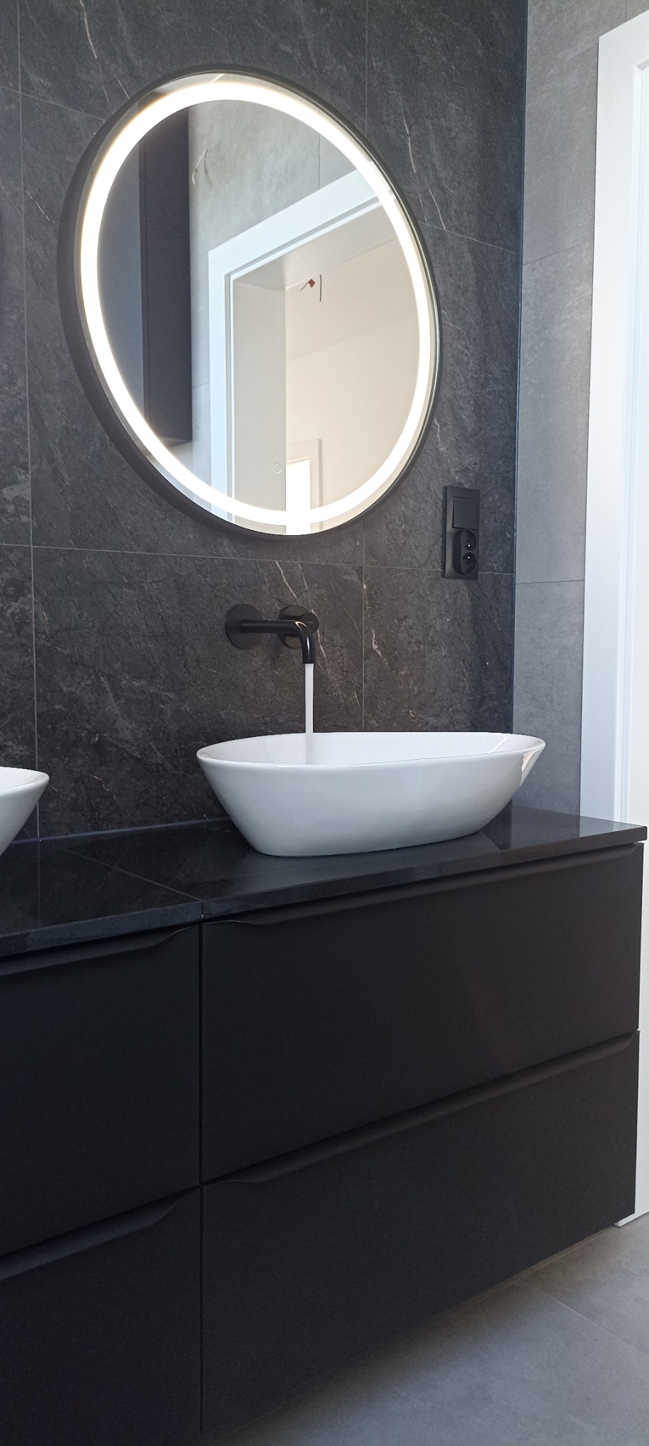 Nowoczesna łazienka z owalną umywalką nablatową, czarną armaturą i okrągłym lustrem z podświetleniem LED na tle ciemnych płytek imitujących marmur.
