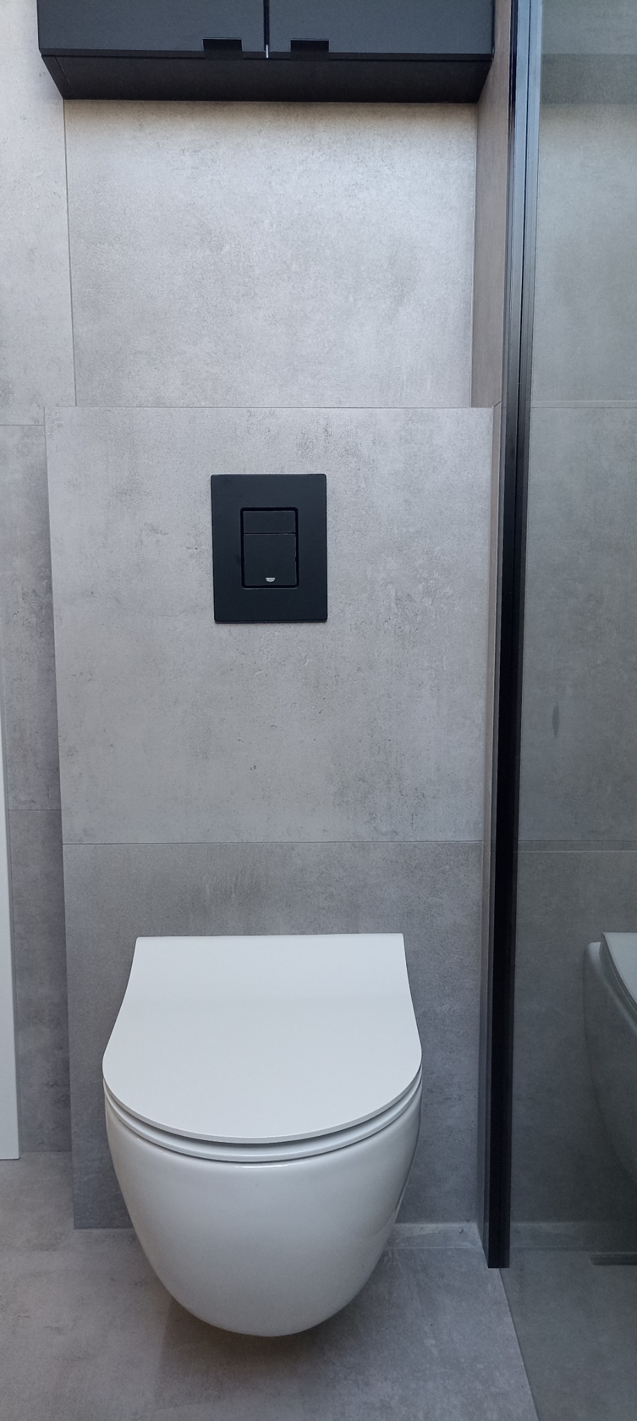 Nowoczesna toaleta podwieszana z czarnym przyciskiem spłukującym na szarej, gładkiej ścianie, częściowo widoczna kabina prysznicowa z czarną ramą.