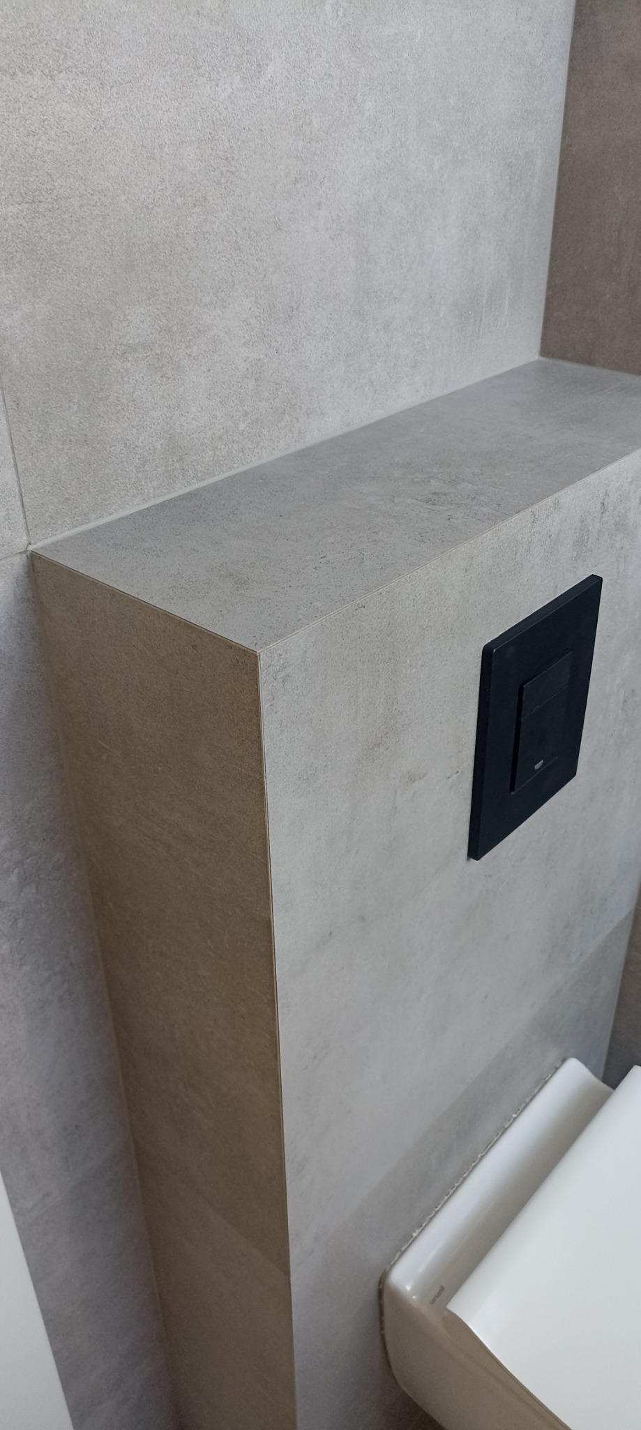 Szara, kamienna zabudowa stelaża WC z czarnym przyciskiem spłukującym, widoczny fragment miski WC.