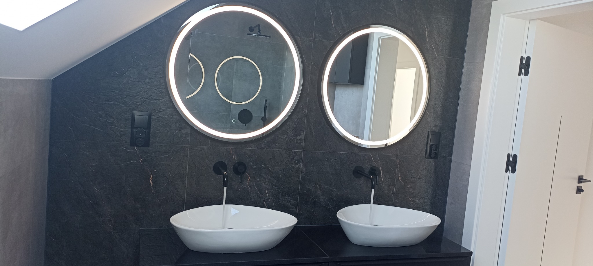Nowoczesna łazienka na poddaszu z dwoma okrągłymi lustrami z podświetleniem LED, czarną armaturą i umywalkami nablatowymi o owalnym kształcie na czarnym blacie.