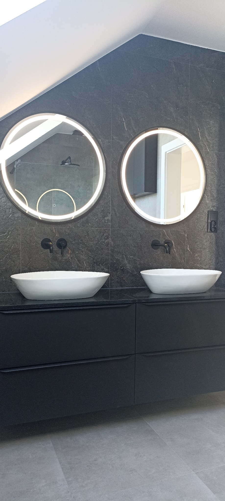 Nowoczesna łazienka z dwoma umywalkami nablatowymi, okrągłymi lustrami z podświetleniem LED i czarną armaturą na tle ciemnych, marmurkowych płytek ściennych i szarej podłogi.