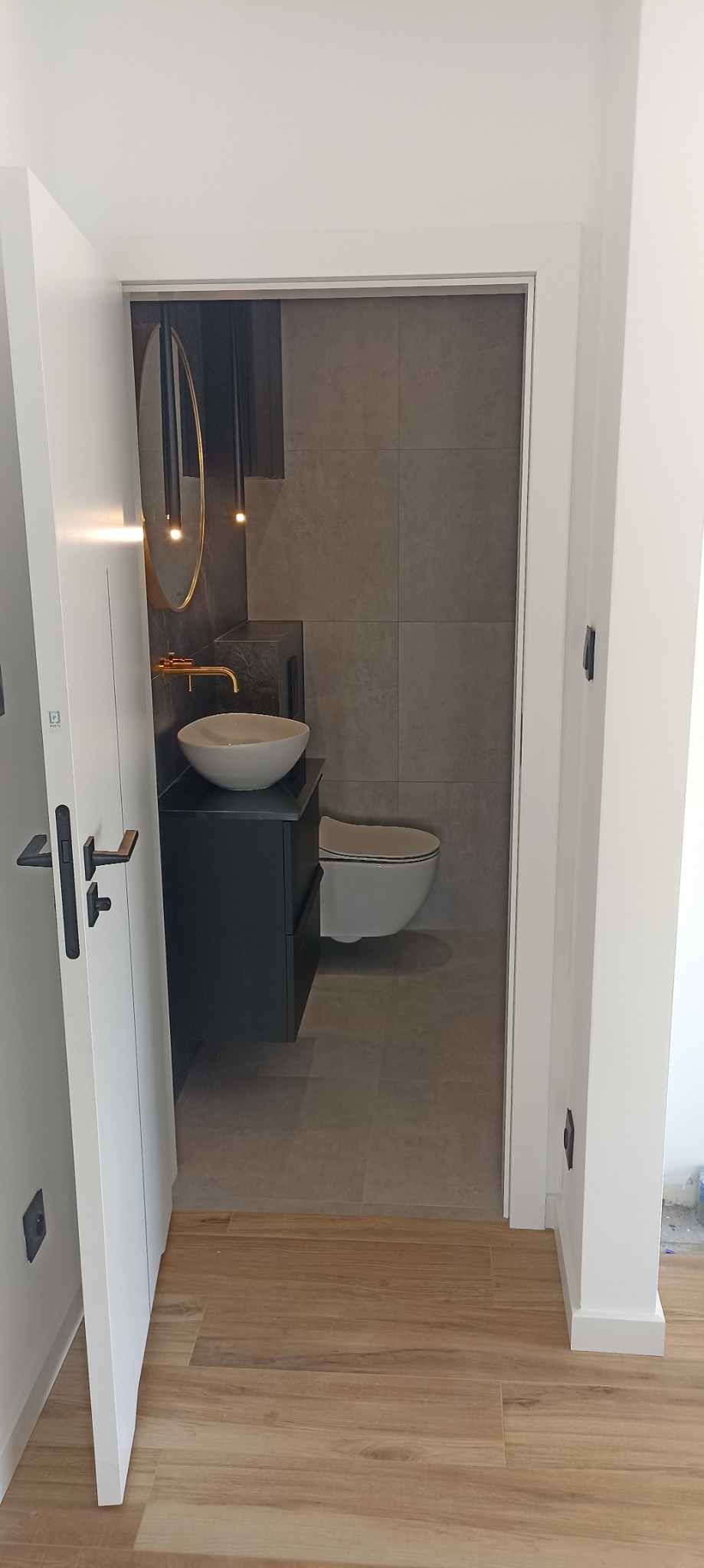 Nowoczesna łazienka z umywalką nablatową na czarnej szafce, złotą armaturą i okrągłym lustrem, widoczna zza otwartych białych drzwi z czarną klamką, podłoga wyłożona panelami imitującymi drewno.