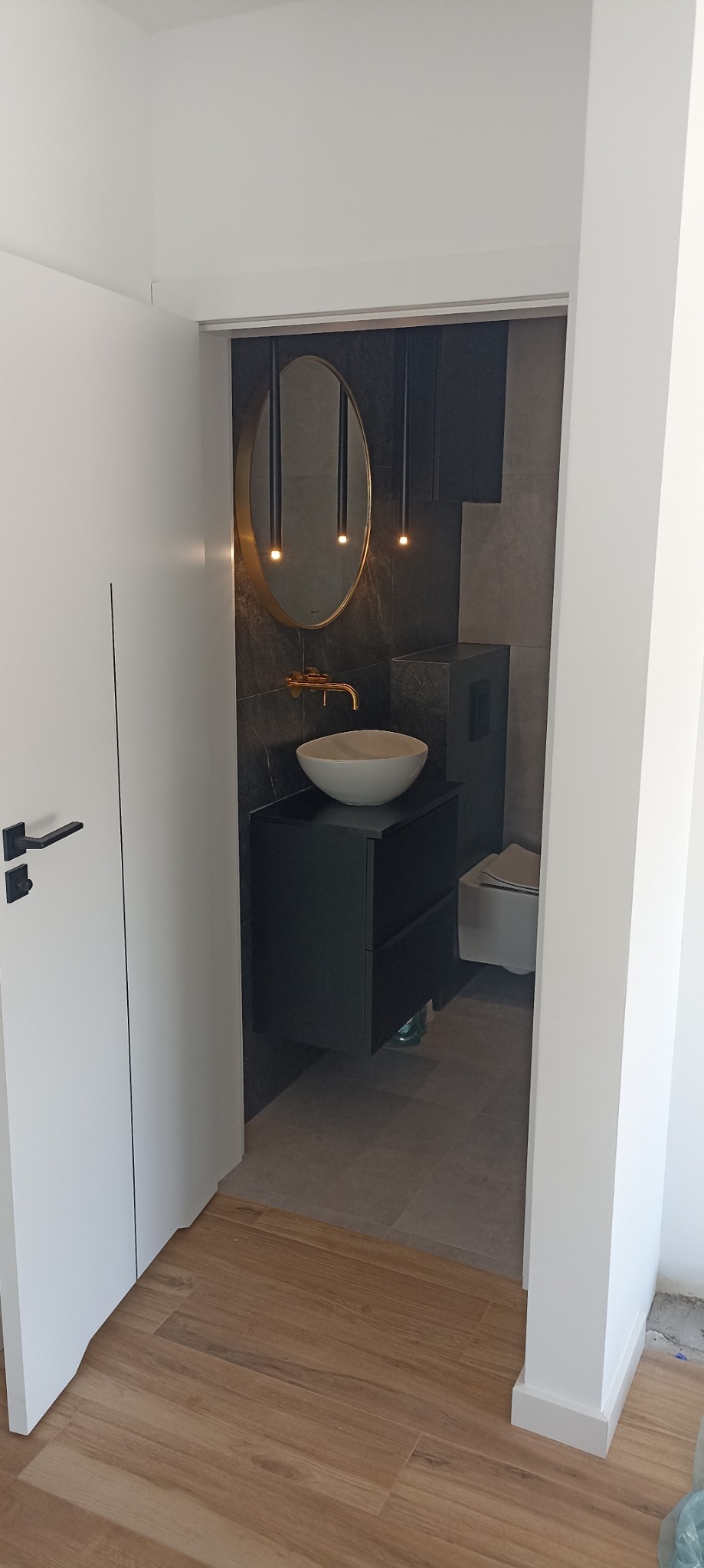 Nowoczesna łazienka z umywalką nablatową, złotą armaturą, owalnym lustrem w złotej ramie i wiszącymi lampami na tle grafitowych płytek, widok z korytarza.