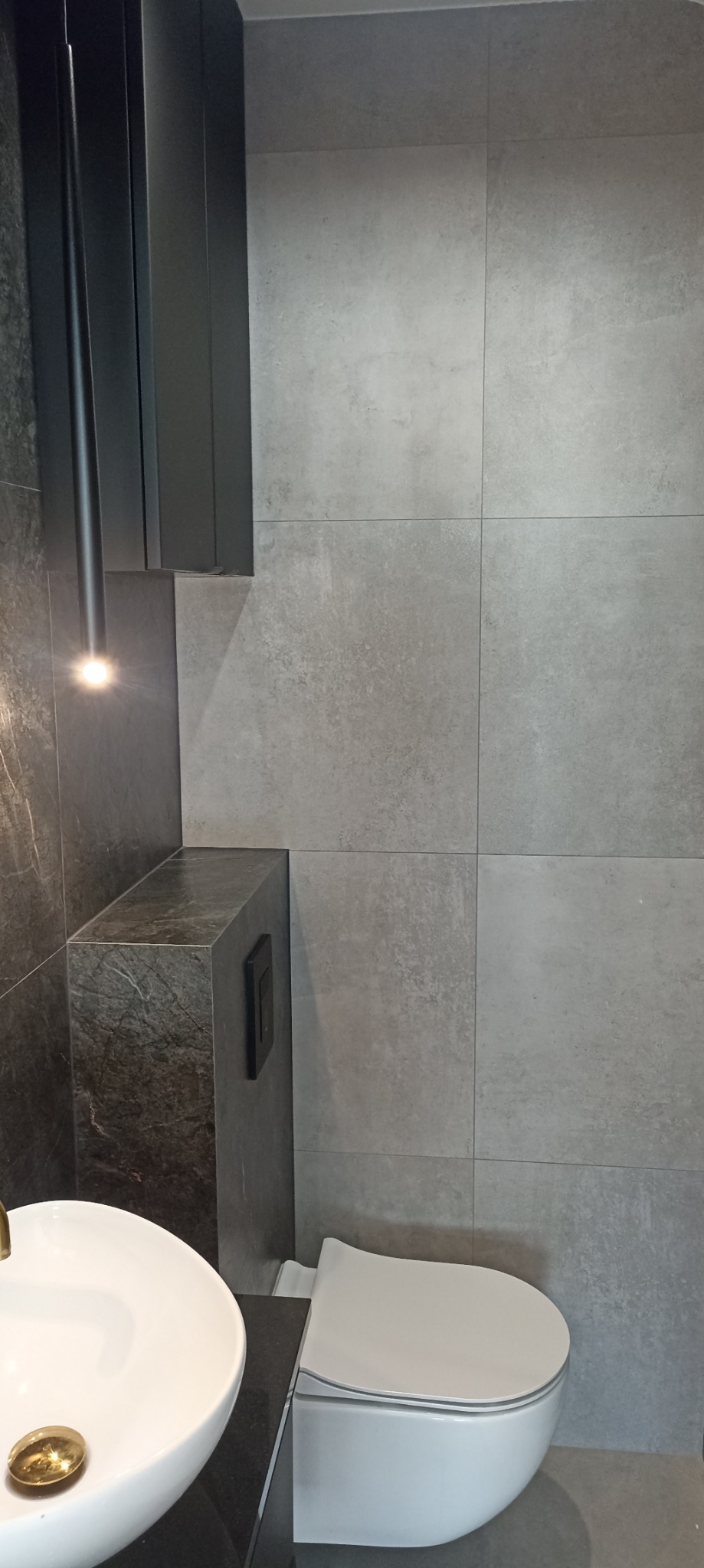 Nowoczesna łazienka z szarą płytką imitującą beton, wisząca toaleta, umywalka nablatowa z czarnym blatem i szafką wiszącą z oświetleniem punktowym.