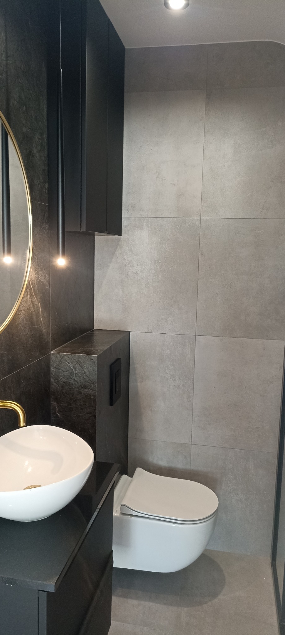 Nowoczesna łazienka z umywalką nablatową na czarnym blacie, podwieszaną toaletą i szarą okładziną ścienną z płytek wielkoformatowych. Lustro w złotej ramie i wiszące lampy nadają elegancji.