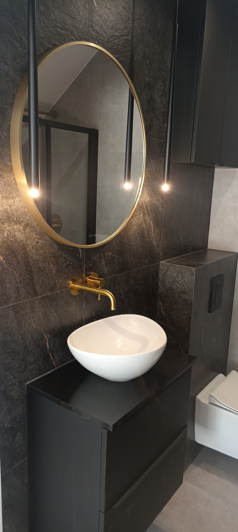 Nowoczesna łazienka z umywalką nablatową, złotą armaturą i okrągłym lustrem podświetlanym wiszącymi lampami, wykończona płytkami imitującymi ciemny marmur.