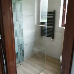 Nowoczesna łazienka z kabiną prysznicową typu walk-in, wykończona płytkami w odcieniach bieli i turkusu, drewnopodobna podłoga, czarny grzejnik drabinkowy i widok na zieleń za oknem.