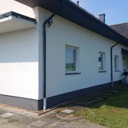 Świeżo pomalowana elewacja domu jednorodzinnego w jasnym kolorze, z widocznymi oknami, ciemną podmurówką i rynnami, na tle błękitnego nieba.