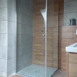 Kabina prysznicowa walk-in z przezroczystego szkła, wykończona szarymi i drewnopodobnymi płytkami, z odpływem liniowym w posadzce wyłożonej mozaiką.