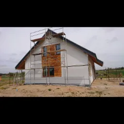 Dom jednorodzinny w trakcie ocieplania ścian, z widocznym rusztowaniem, częściowo otynkowaną elewacją w kolorze złamanej bieli oraz drewnianą okładziną elewacyjną wokół okien na poddaszu.