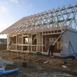 Konstrukcja szkieletowa domu jednorodzinnego w trakcie budowy, widoczne drewniane elementy nośne i więźba dachowa, pracownik w tle, teren budowy z materiałami i narzędziami.
