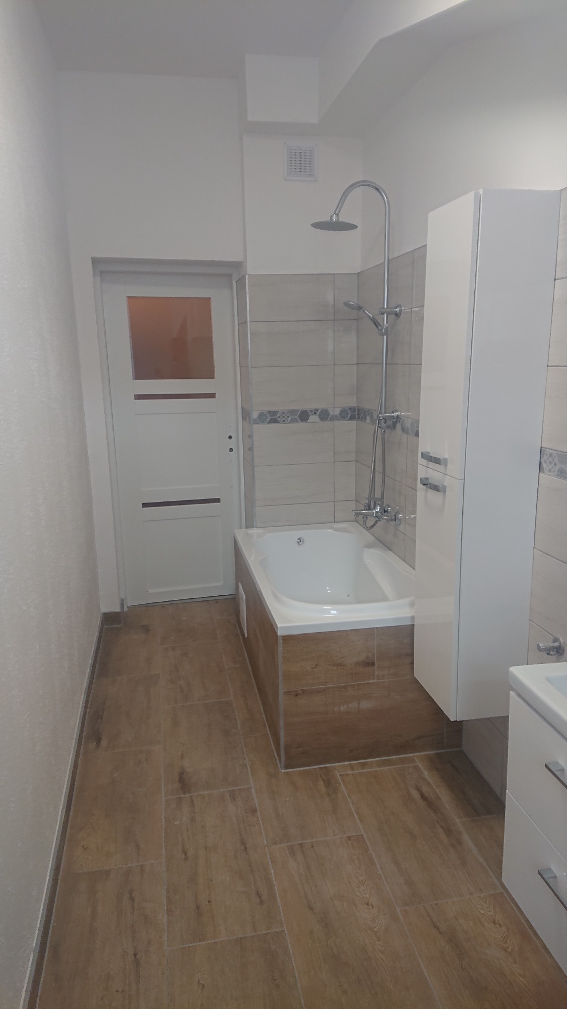 Wykończona łazienka z wanną obudowaną płytkami imitującymi drewno, kabiną prysznicową, białą szafką łazienkową i podłogą z płytek imitujących drewno, widok na białe drzwi z szybą.