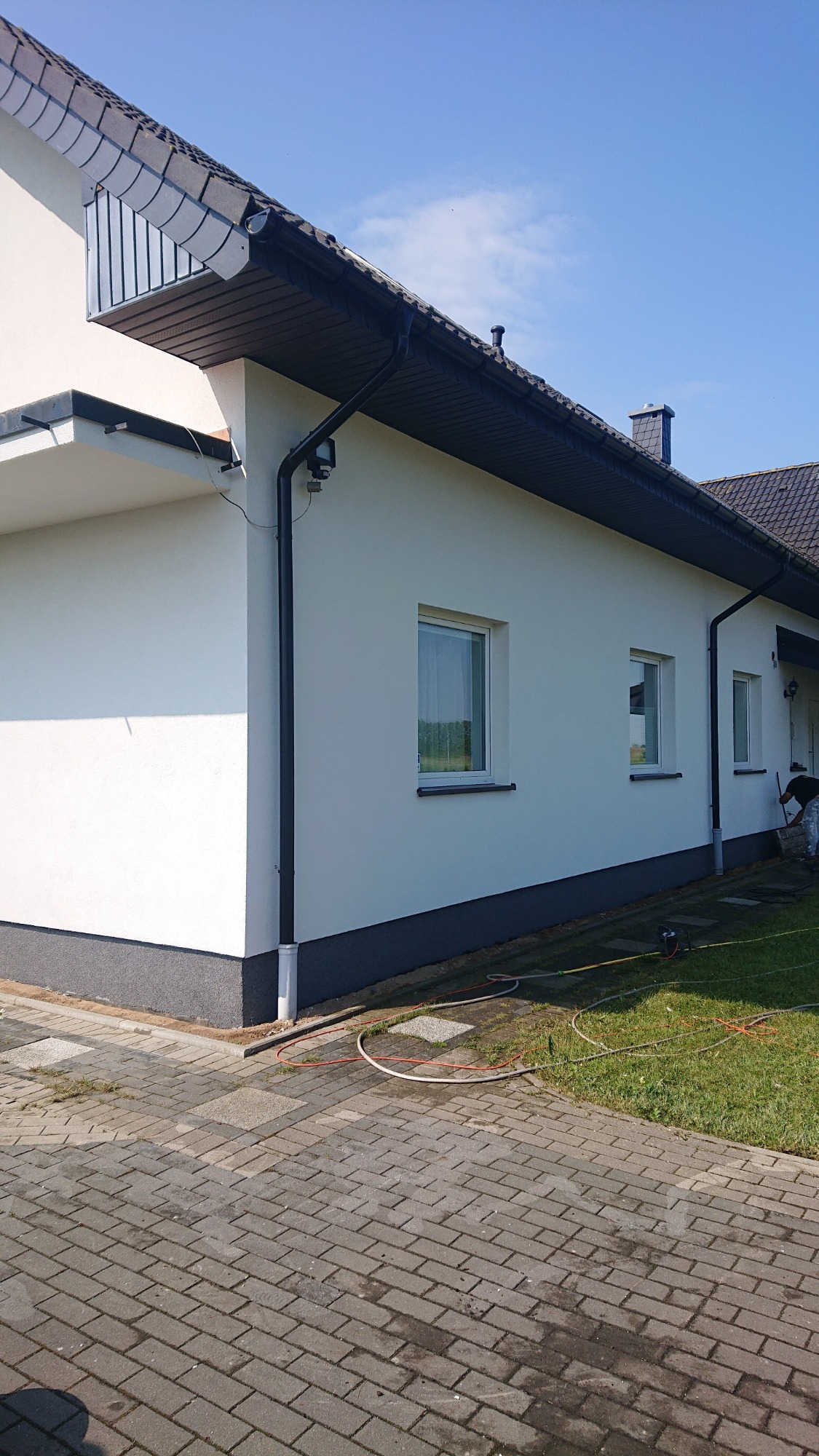 Świeżo pomalowana elewacja domu jednorodzinnego w jasnym kolorze, z widocznymi oknami, ciemną podmurówką i rynnami, na tle błękitnego nieba.