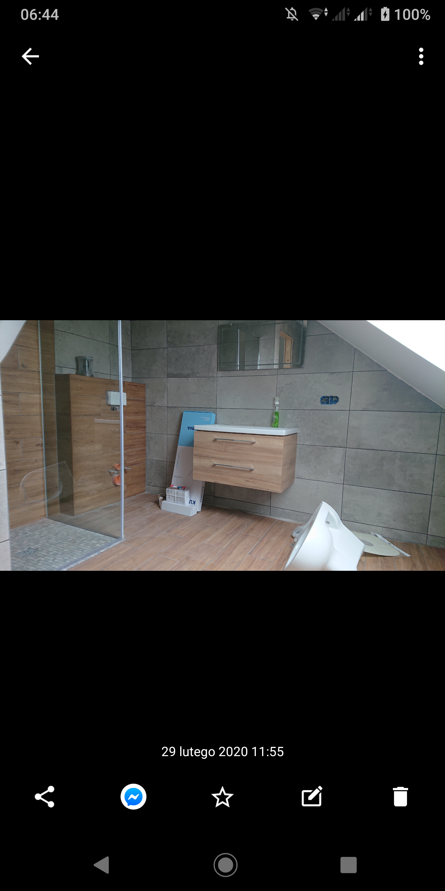 Niedokończona łazienka na poddaszu z kabiną prysznicową typu walk-in, umywalką z szafką wiszącą i częściowo zamontowaną toaletą; widoczne płytki na ścianach i podłodze, kartony po produktach.