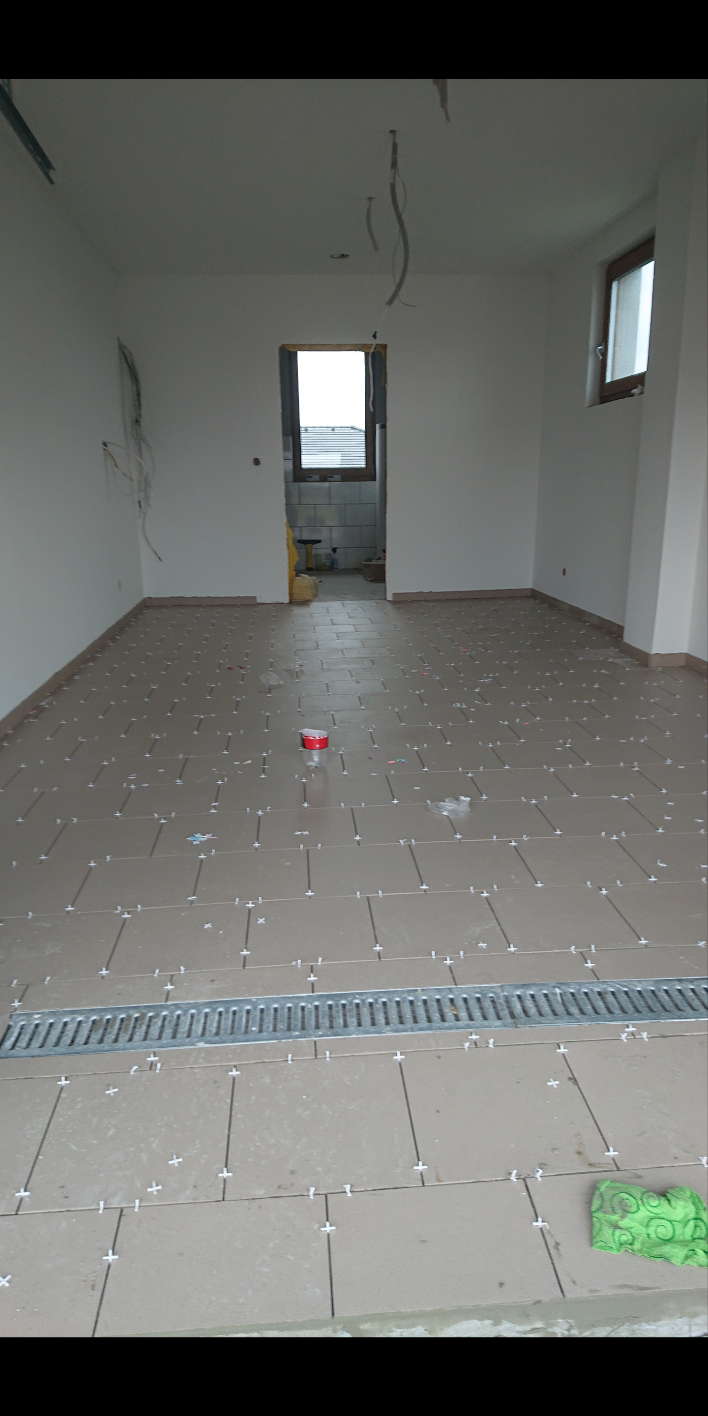 Układanie płytek podłogowych w jasnym pomieszczeniu z widocznym odpływem liniowym, krzyżyki dystansowe i resztki zaprawy, w tle otwarte drzwi do łazienki z białymi płytkami.