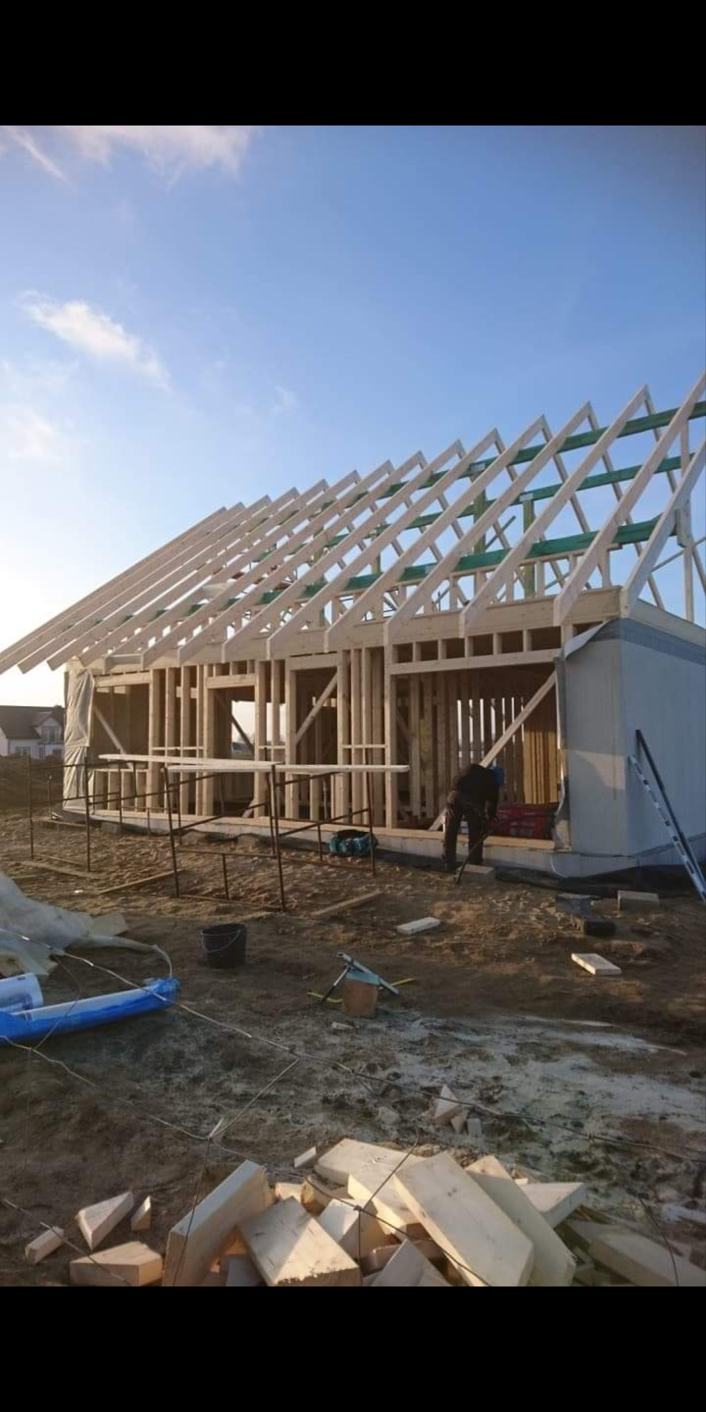 Konstrukcja szkieletowa domu jednorodzinnego w trakcie budowy, widoczne drewniane elementy nośne i więźba dachowa, pracownik w tle, teren budowy z materiałami i narzędziami.