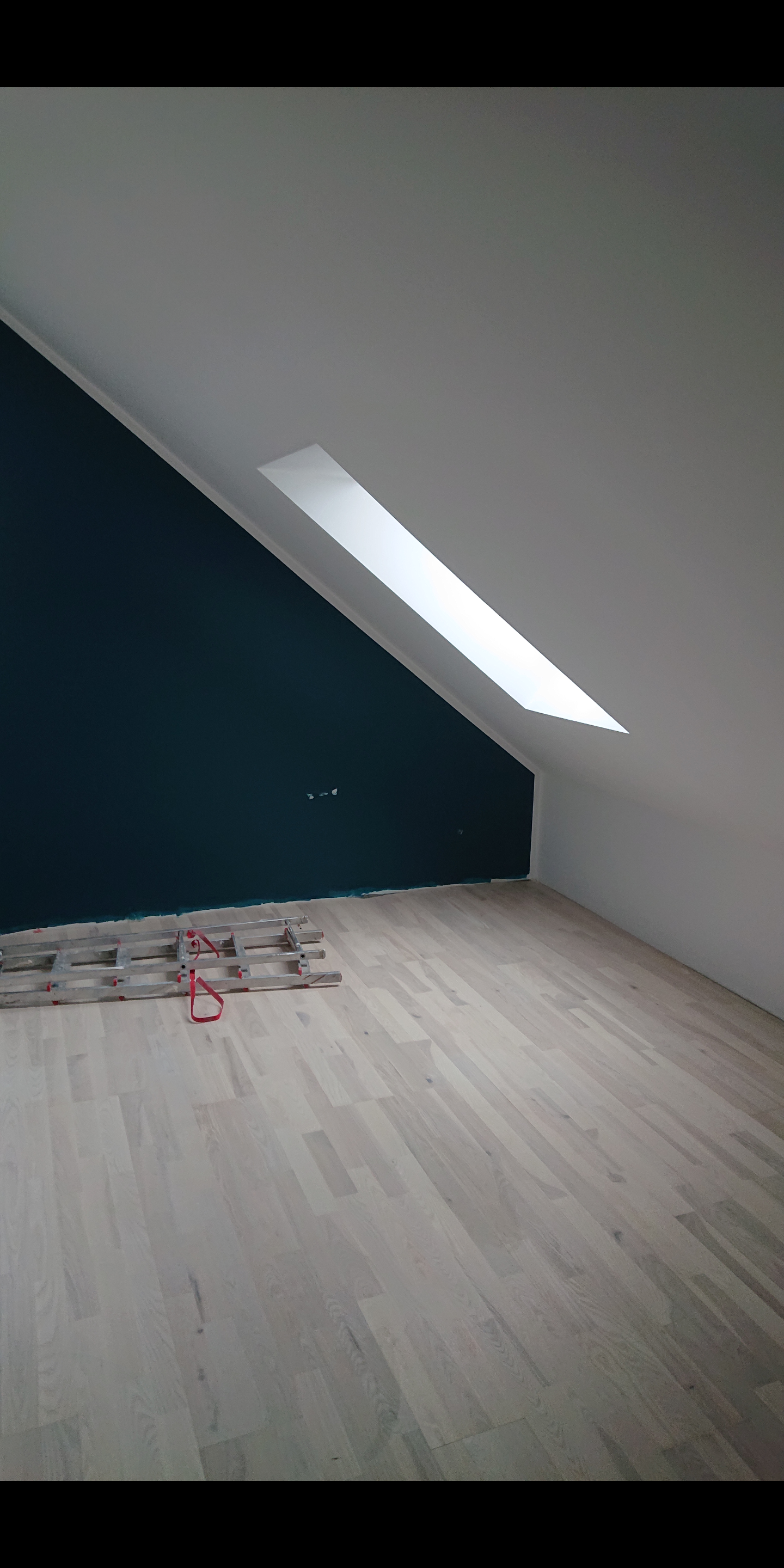 Pomieszczenie na poddaszu po malowaniu: ściana pomalowana na ciemny turkus, biały sufit ze świetlikiem, parkiet z jasnego drewna, rozłożona aluminiowa drabina malarska.
