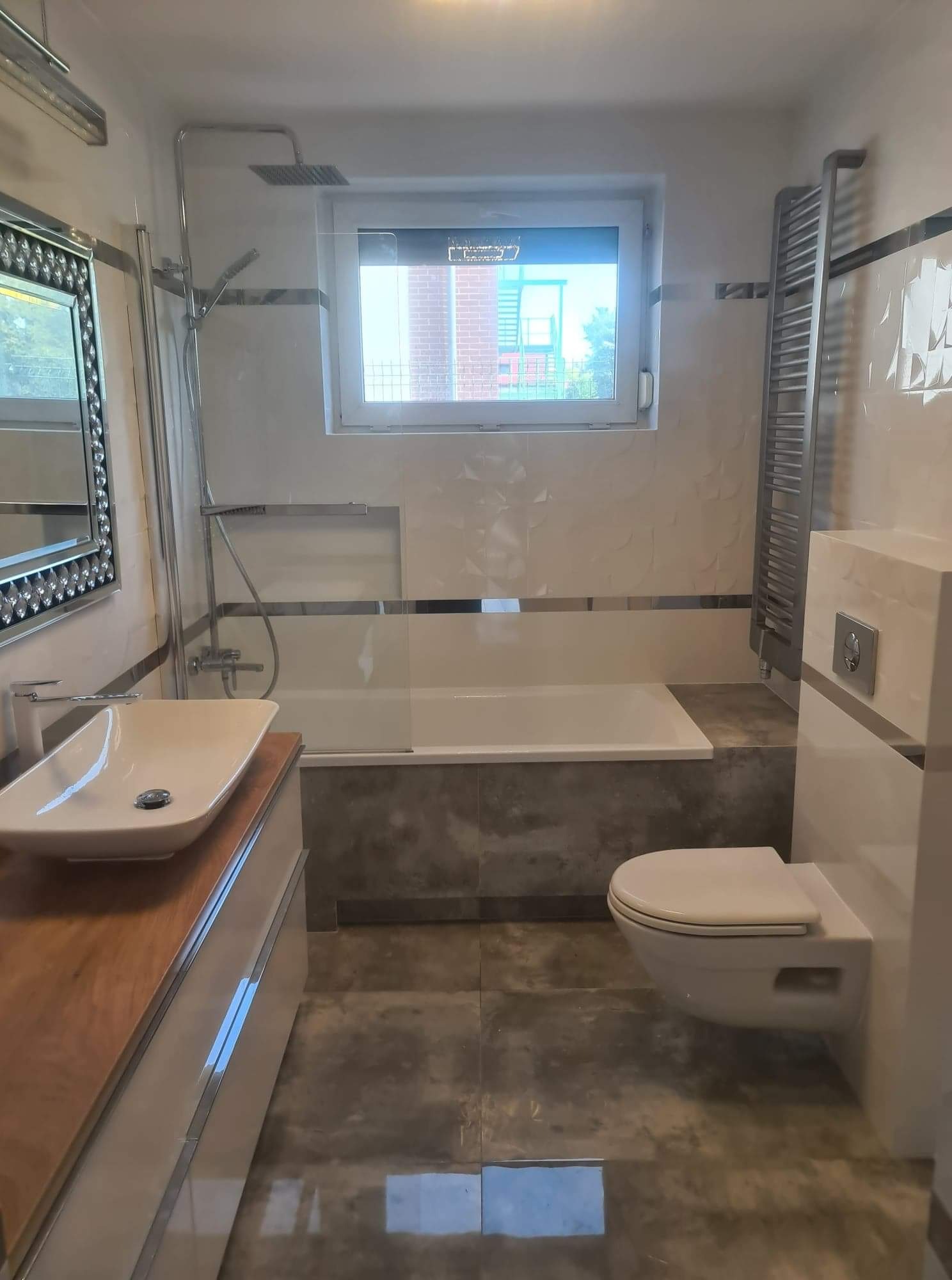 Nowoczesna łazienka z wanną, umywalką nablatową na drewnianym blacie i podwieszaną toaletą, wykończona płytkami w odcieniach bieli i szarości z dekoracyjnym lustrem.