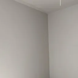 Świeżo otynkowany, gładki narożnik pokoju w odcieniach szarości, z widocznym fragmentem sufitu i wiszącym kablem.