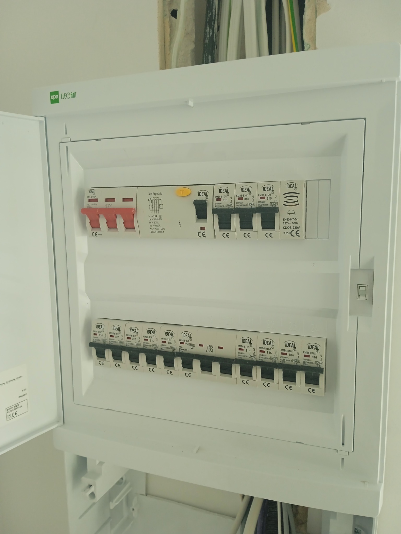 Otwarta skrzynka rozdzielcza z bezpiecznikami, widoczne kable w ścianie, bezpieczniki marki Ideal, etykieta EPN Elegant na górze.