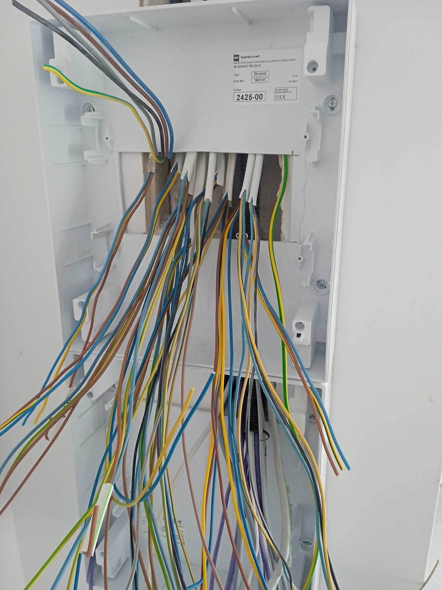 Otwarta skrzynka elektryczna z wystającymi kablami w różnych kolorach, częściowo w ścianie, widoczne oznaczenia producenta na obudowie.