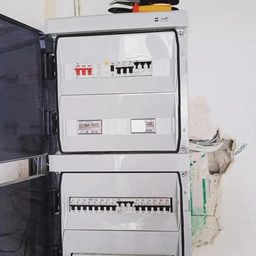 Otwarta skrzynka elektryczna z licznikami i bezpiecznikami, widoczna instalacja elektryczna w ścianie, narzędzia na górze obudowy, ściana z ubytkiem tynku.