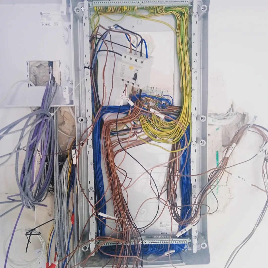 Rozdzielnia elektryczna w trakcie montażu, widoczne liczne przewody w różnych kolorach, listwy zaciskowe i zabezpieczenia, ściana z widocznymi śladami po kuciu.